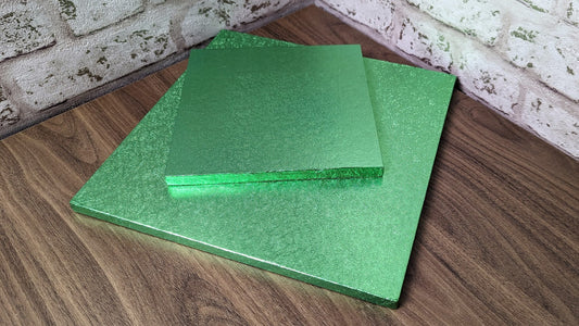 Two green square cake drums made of compressed cardboard with a colored foil covering, placed one on top of the other on a wooden surface.