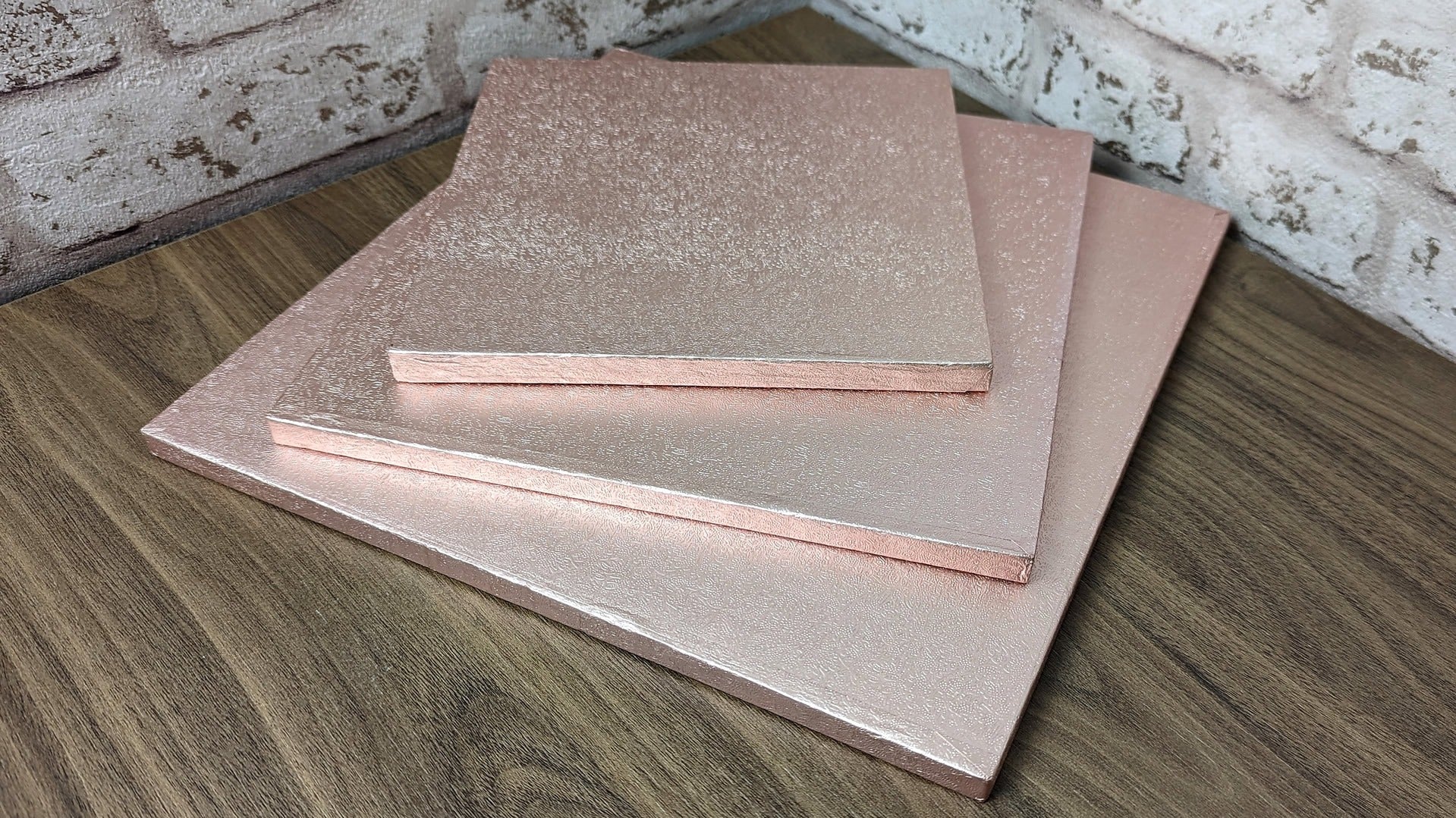 A stack of rose gold colored square cake drums made of compressed cardboard with a colored foil covering.