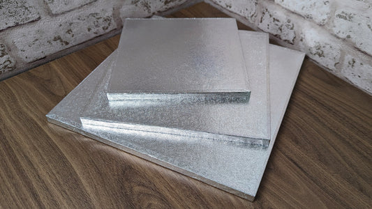 A stack of square silver cake drums made of cardboard on a wooden surface.